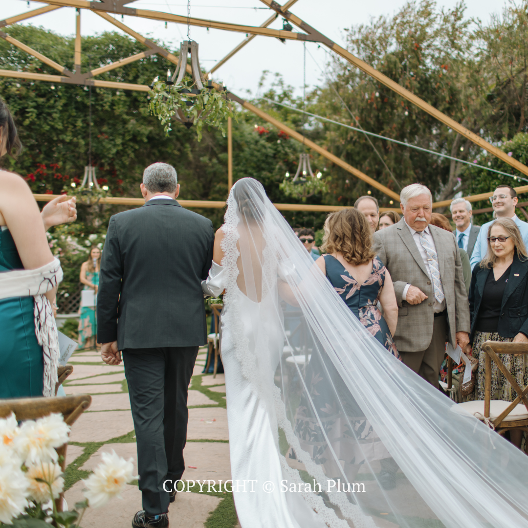 Wedding guests walking down a garden path under an arch, with the bride in a long veil and train.