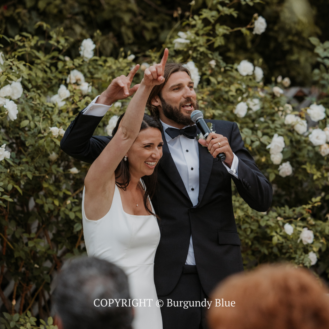 Couple in formalwear celebrating outdoors, smiling and raising their hands with microphones in front of flowers