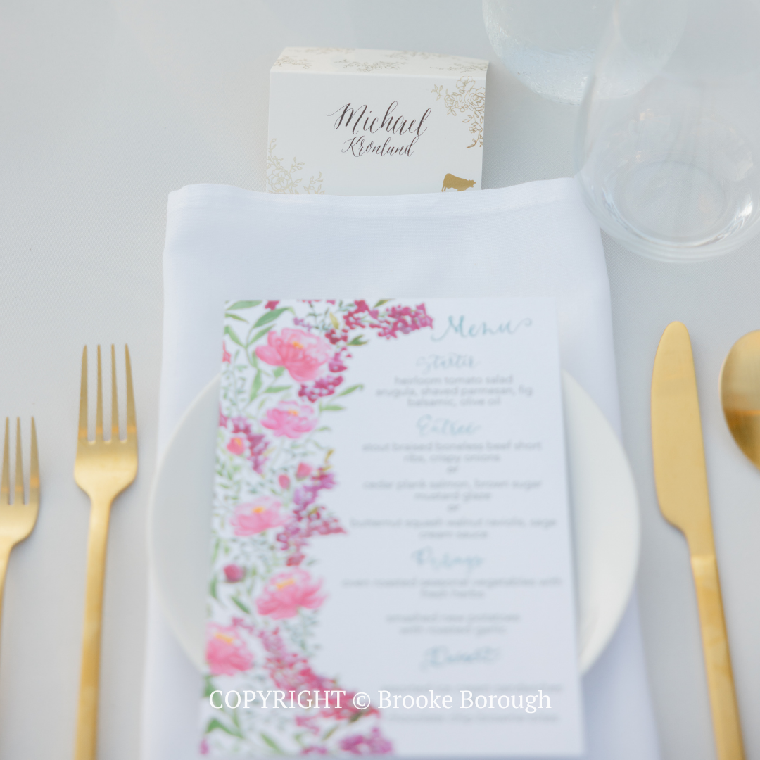 White wedding menu with pink floral border on a white plate, set with gold utensils on a light table setting