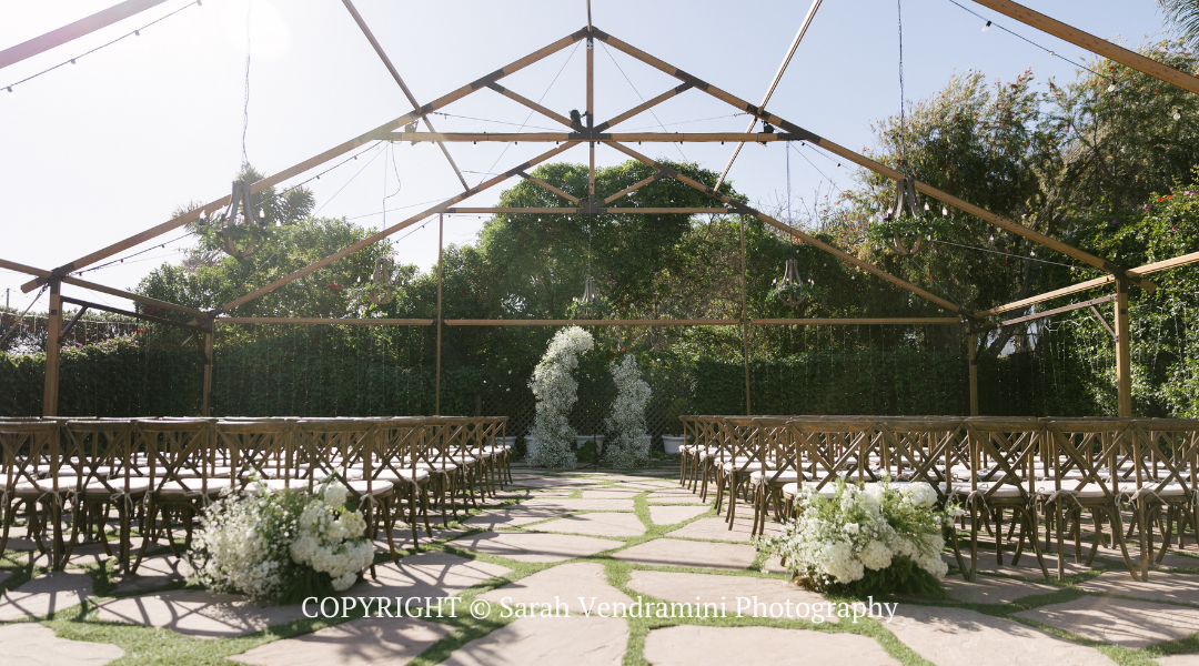 Outdoor wedding ceremony setup with white chairs, floral aisle, and glass-roofed greenhouse backdrop