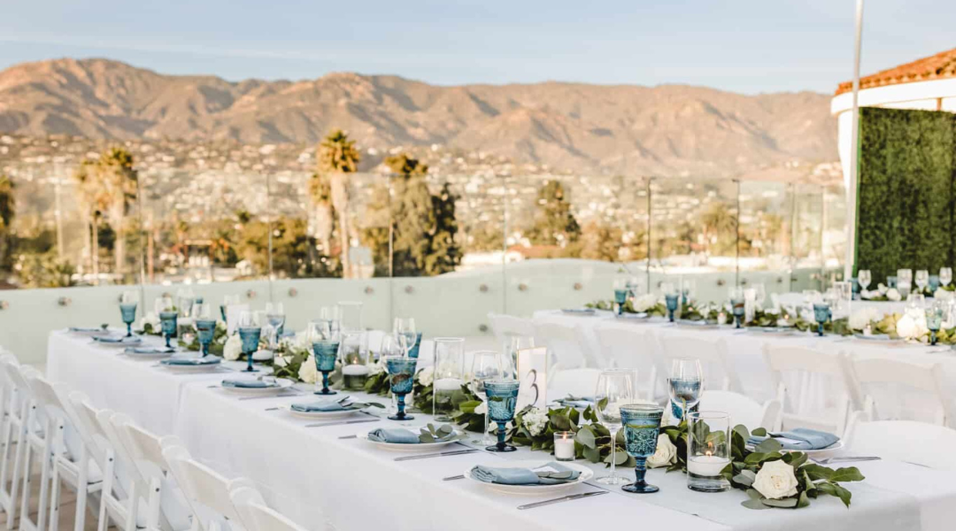 Outdoor banquet table set with white linens and floral centerpieces, mountains in the background