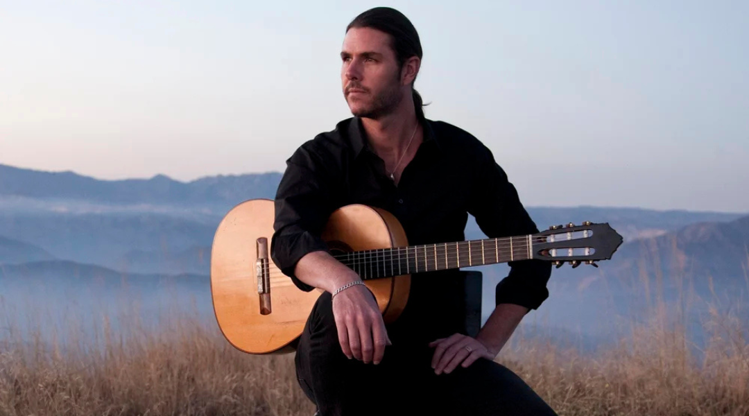 Man seated outdoors at dusk holding an acoustic guitar, with mountains in the background