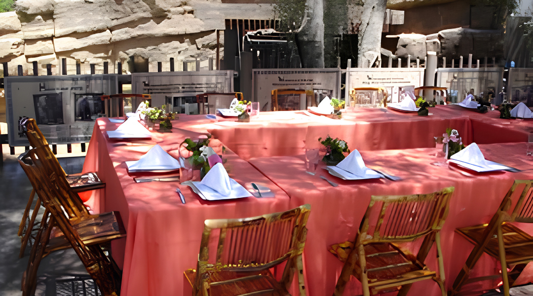 Outdoor restaurant table set with red tablecloth, white folded napkins, and bamboo chairs in a courtyard.