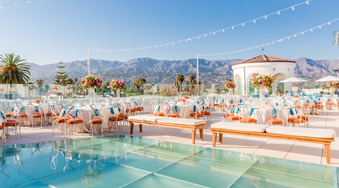 Sunlit outdoor wedding reception by a pool, with white chairs, floral decor, and mountains in the background