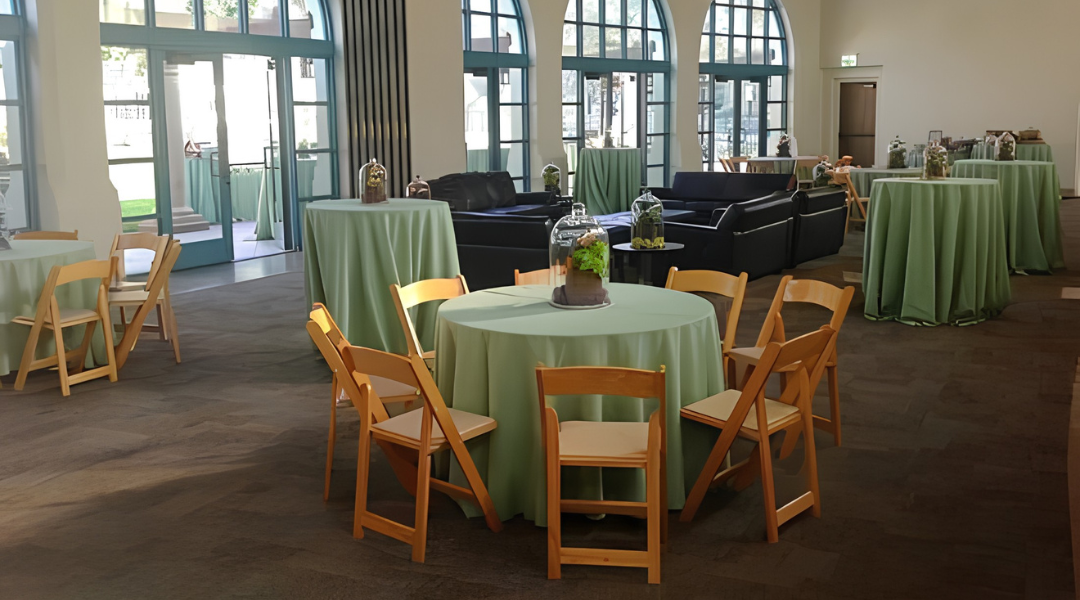 Indoor banquet hall with round tables draped in green tablecloths and wooden chairs near large windows