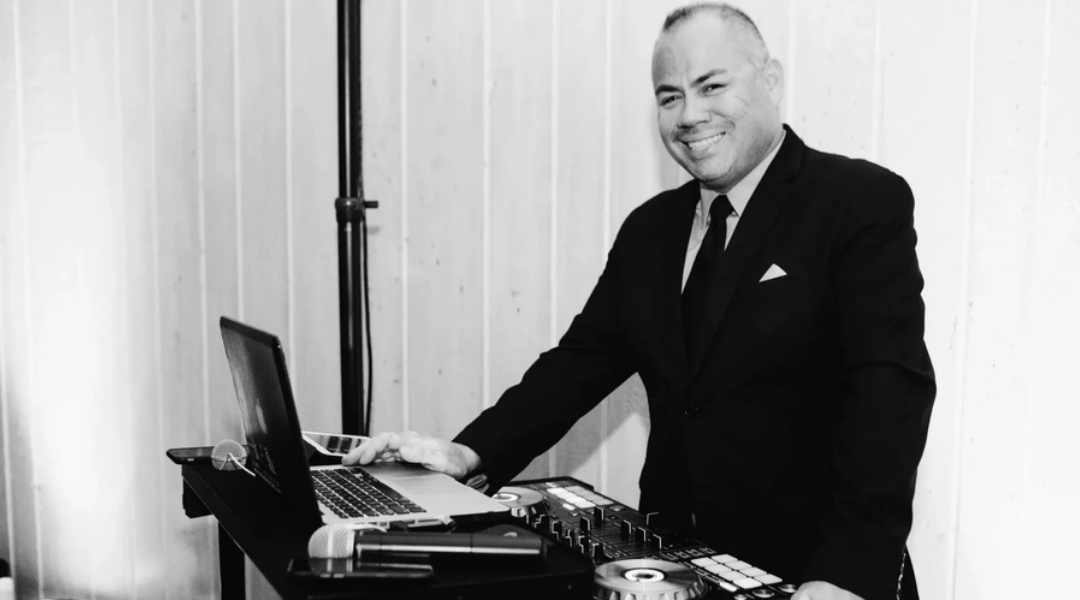 Man in a dark suit smiling beside a laptop on a table, indoors