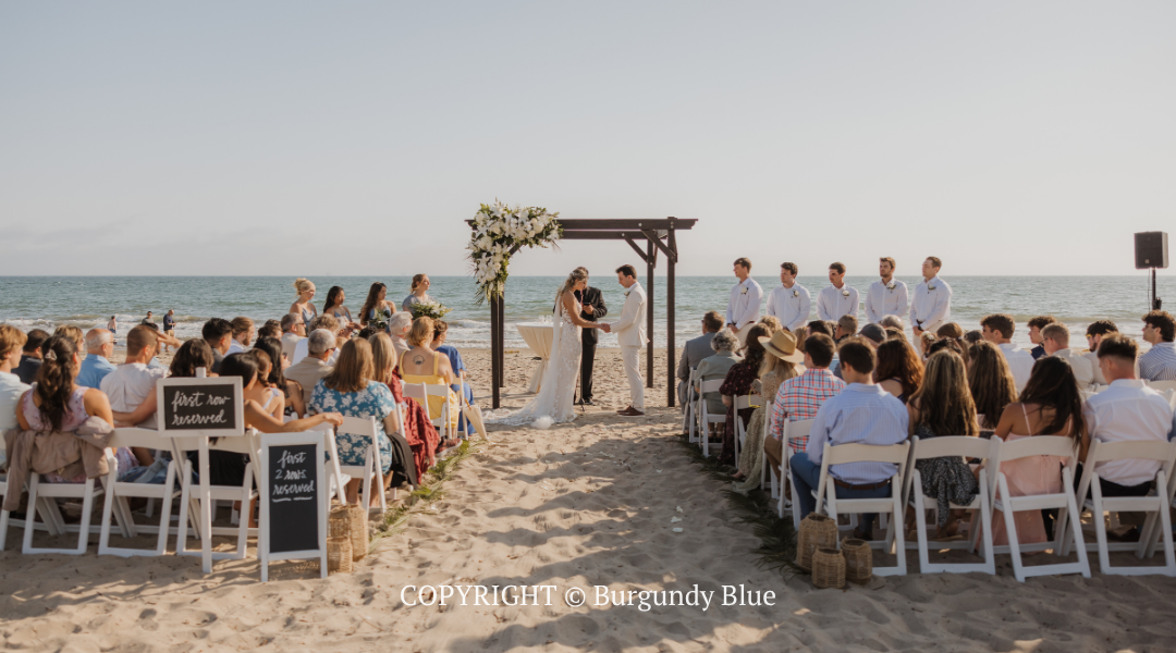 Beach wedding ceremony with guests seated on chairs under a decorated arch by the ocean