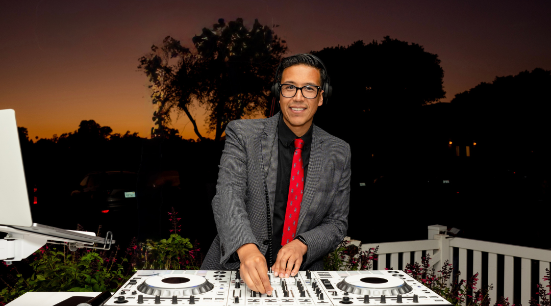 DJ smiling behind a mixer at dusk, wearing a gray jacket and red tie, with outdoor lights in the background