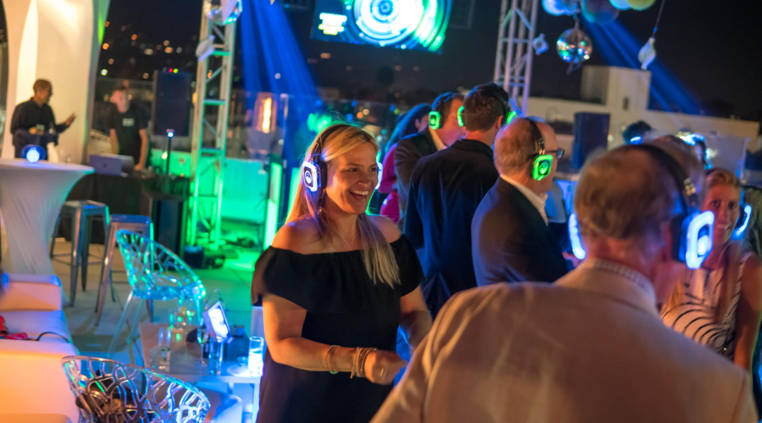 People wearing glowing headphones dance at a lively indoor party with blue and green lights.