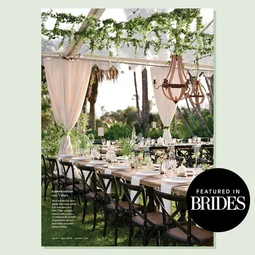 Elegant outdoor wedding reception under a draped canopy with long table settings, greenery, and chandeliers.