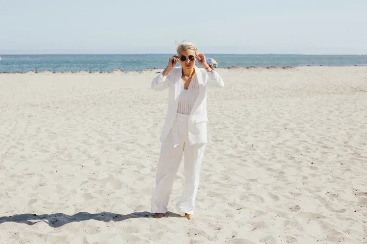 Person in white suit standing on a sandy beach, holding binoculars with the ocean in the background