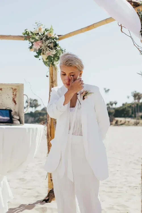 Woman in white suit and veil standing on a sandy beach under a floral arch
