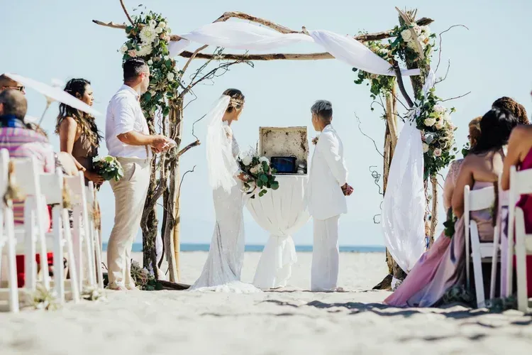 Beach wedding ceremony under a floral arch with guests seated on both sides