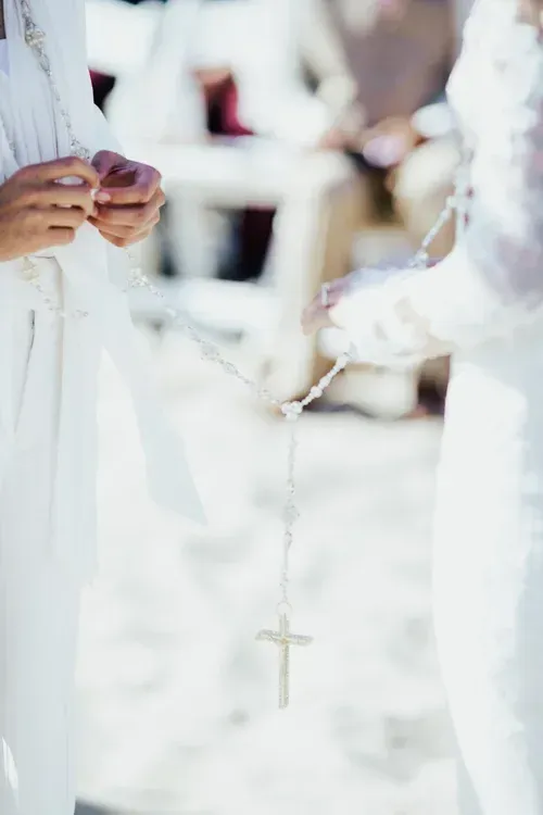 Hands holding rosary beads in a white-lit church setting, with a cross necklace visible.