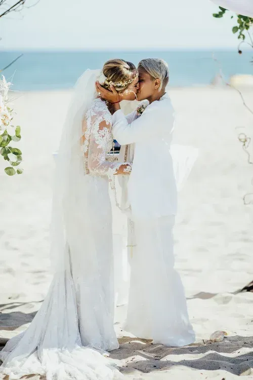 Two brides in white gowns and veils embrace on a sunny beach by the ocean.