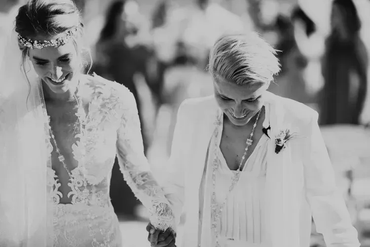 Two smiling women in white dresses holding hands outdoors at a wedding ceremony