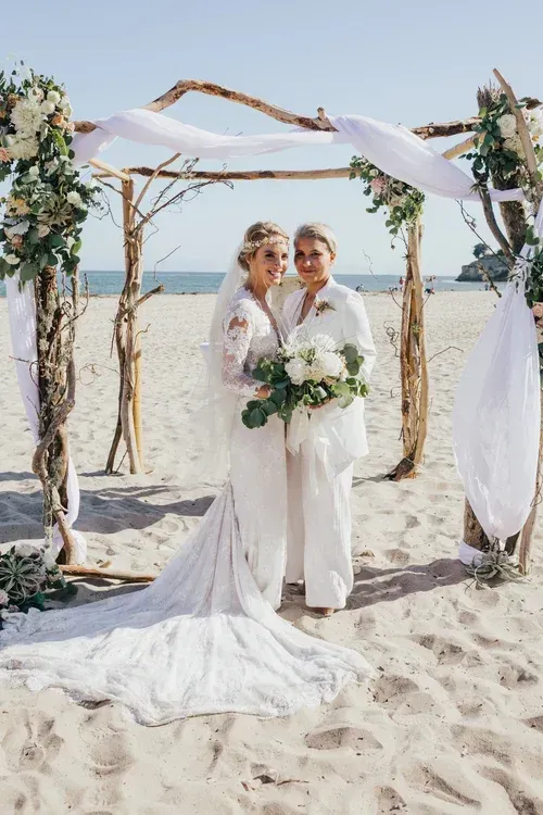Wedding couple standing under a rustic arch on a beach, holding bouquets with long white fabric draping overhead