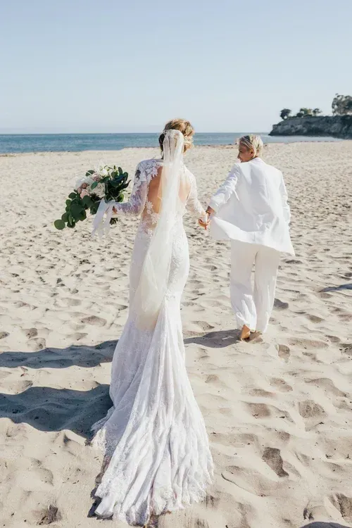 Bride and partner walking on a sandy beach, holding flowers, with ocean in the background
