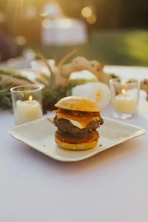 Mini burger on a square plate with candles and a blurred outdoor dining table in the background