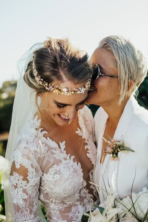 Two women in white wedding attire, one kissing the other on the forehead outdoors.