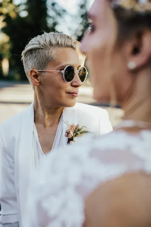 Person in sunglasses and white jacket smiles at a blurred figure in a bright outdoor setting