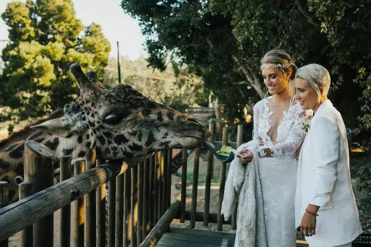 Two women in white outfits feed a giraffe through a wooden fence at an outdoor zoo exhibit