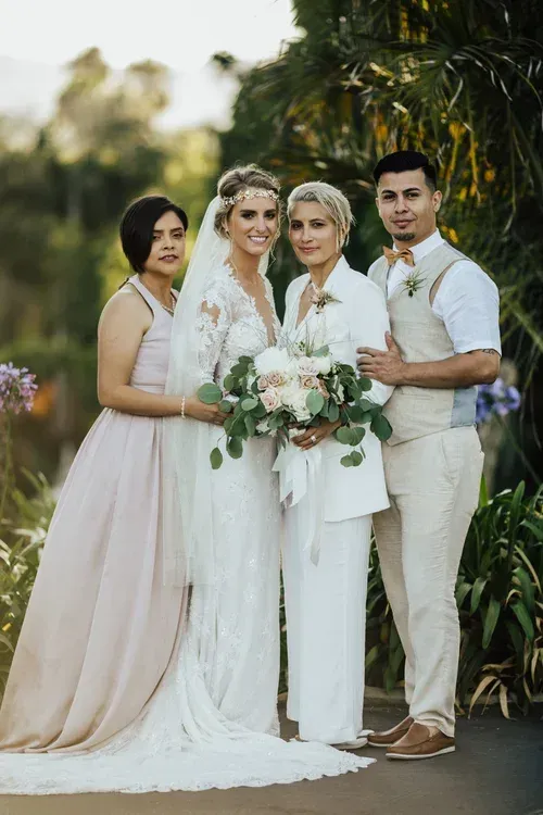 Four people in white formalwear pose outdoors, holding a bouquet at a wedding ceremony.