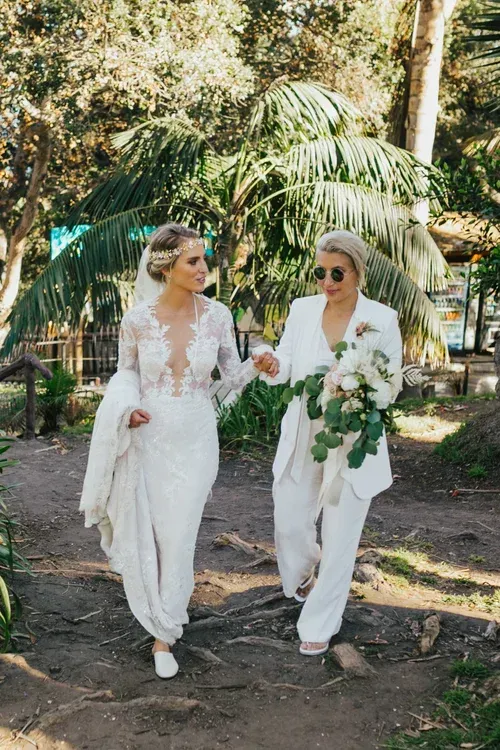 Two brides in white suits walk hand in hand in a sunny garden, each holding a bouquet.