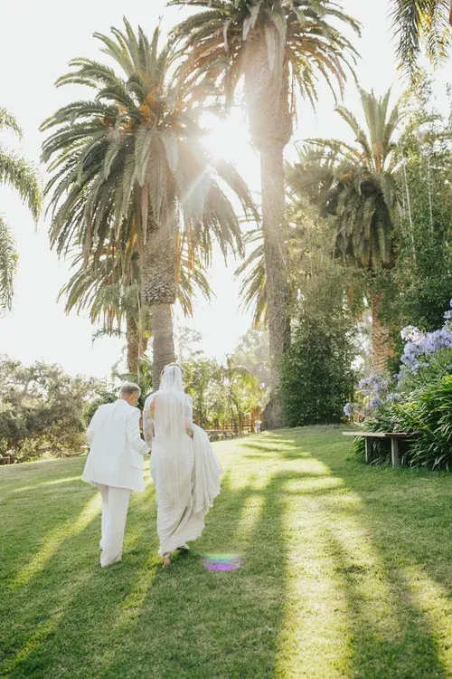 Two people in light clothing walking through a sunny palm garden on a grassy lawn