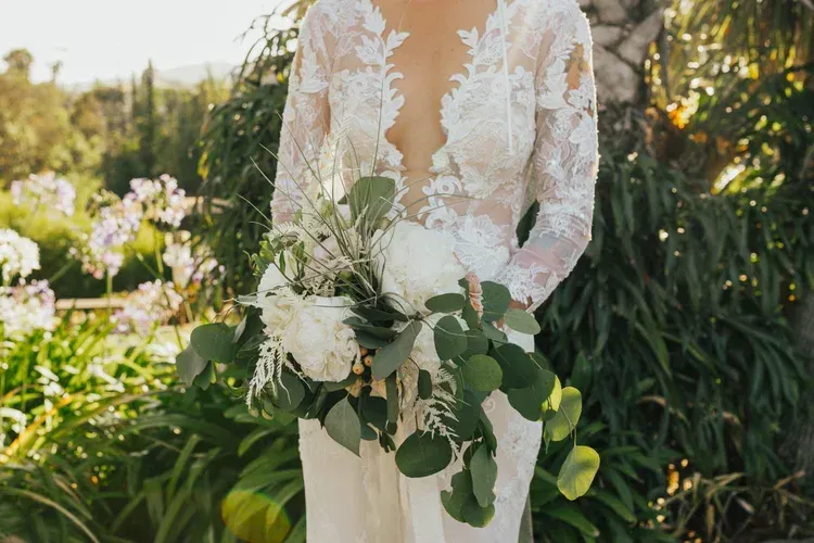 Person holding a white floral bouquet in a sunlit garden, wearing a lace dress