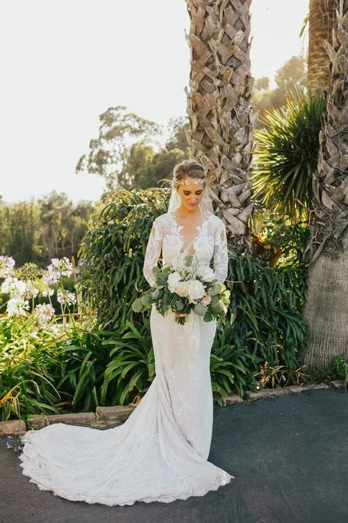 Bride in a white lace gown holding a bouquet, standing outdoors by a palm tree and greenery.