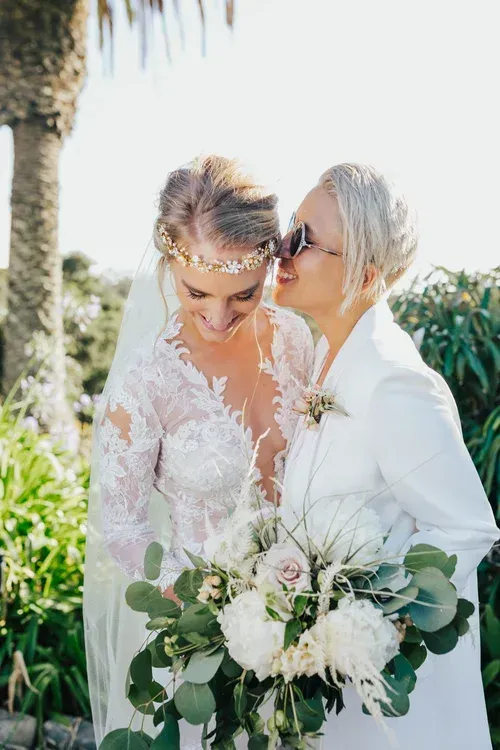 Two women in white outfits embracing at an outdoor wedding, with flowers and greenery nearby.