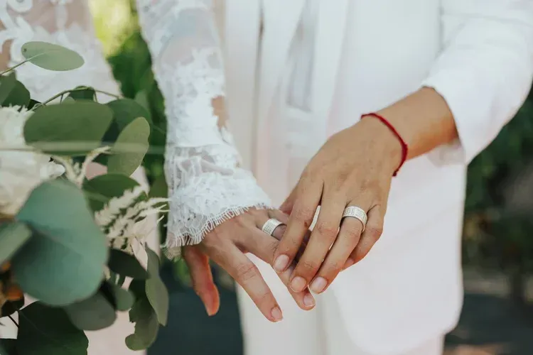 Two hands with wedding rings touching beside a white bouquet and greenery, with a person in white in the background