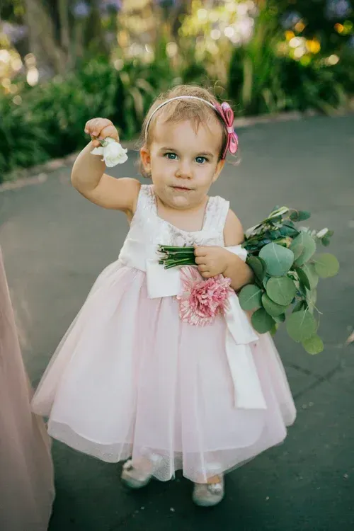 Toddler in a pink dress holding flowers and a bouquet on a sidewalk outdoors