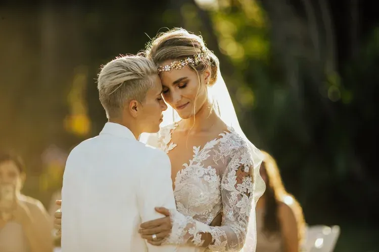 Two people in white wedding attire embrace outdoors in warm sunlight, with a soft blurred background.