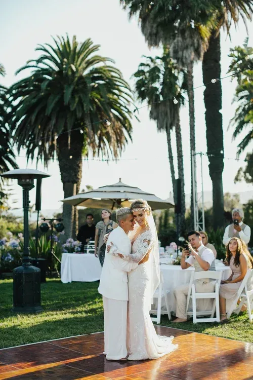 Couple in white embraces at an outdoor wedding reception with palm trees and seated guests.