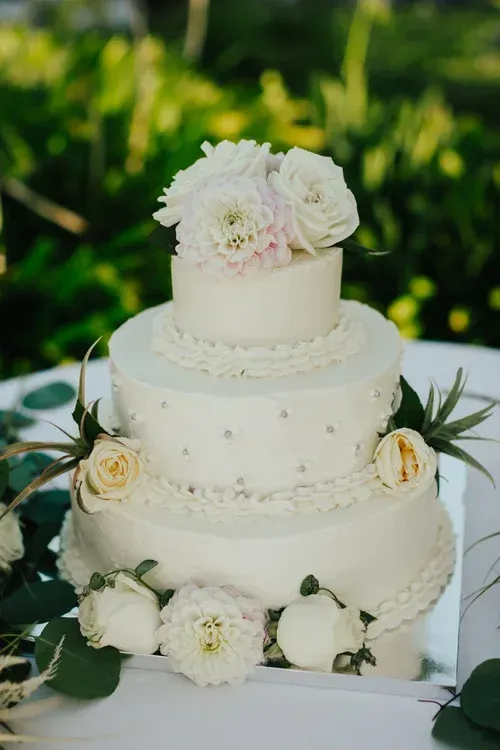 Three-tier white wedding cake with white flowers and greenery on a decorative table