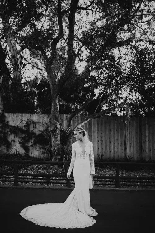 Person in a white gown standing outdoors beneath a large tree, with a fence in the background.
