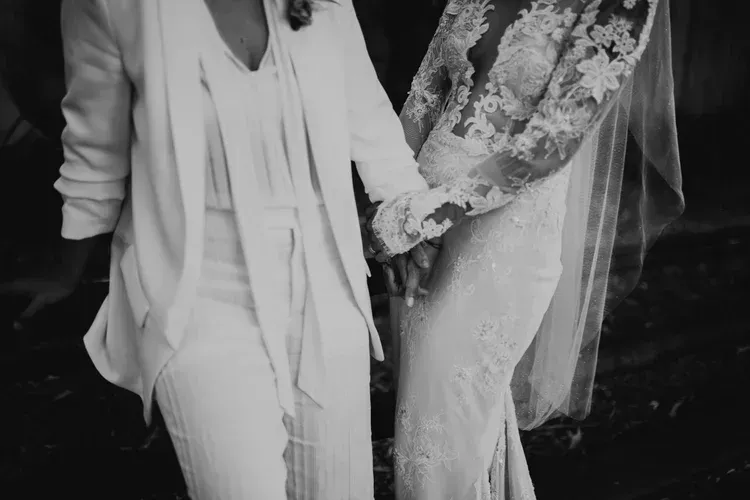 Two people in formal attire holding hands, one in a suit and one in a floral lace dress and veil.