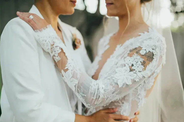 Two people in white lace wedding attire embracing outdoors, one wearing a veil