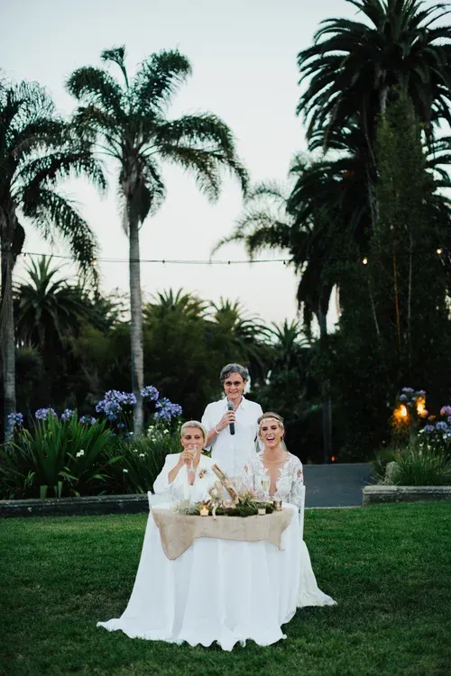 Three people in white seated at a table in a garden at dusk, with palm trees in the background