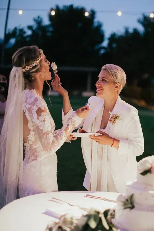 Two people in white dresses share a toast outdoors at dusk, with a candlelit table in the foreground.