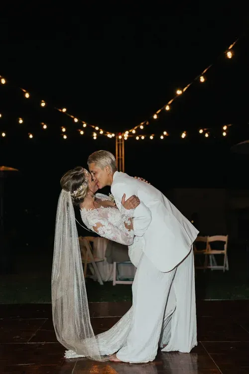Couple kissing at night under string lights, the bride in a long white veil and the groom in a white suit.