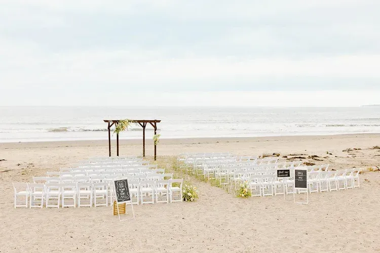Beach wedding setup with white chairs facing a small arch near the shoreline at sunrise