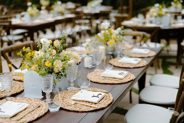 Outdoor banquet table set with woven placemats, white napkins, glassware, and small flower arrangements
