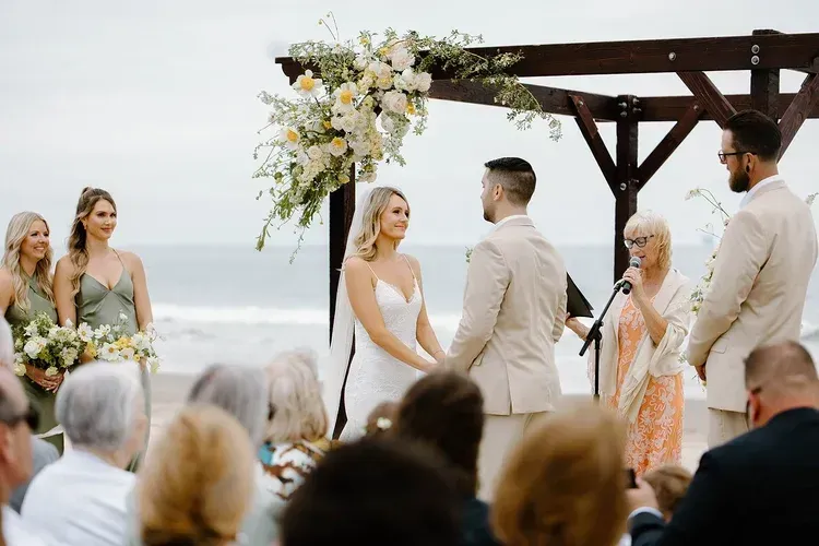 Beach wedding ceremony under floral arch with couple facing each other and guests seated nearby