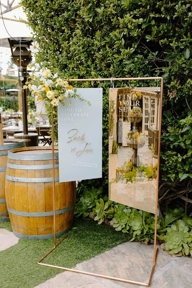 Gold-framed floral display with white and gold signs beside a wooden barrel and hedge backdrop