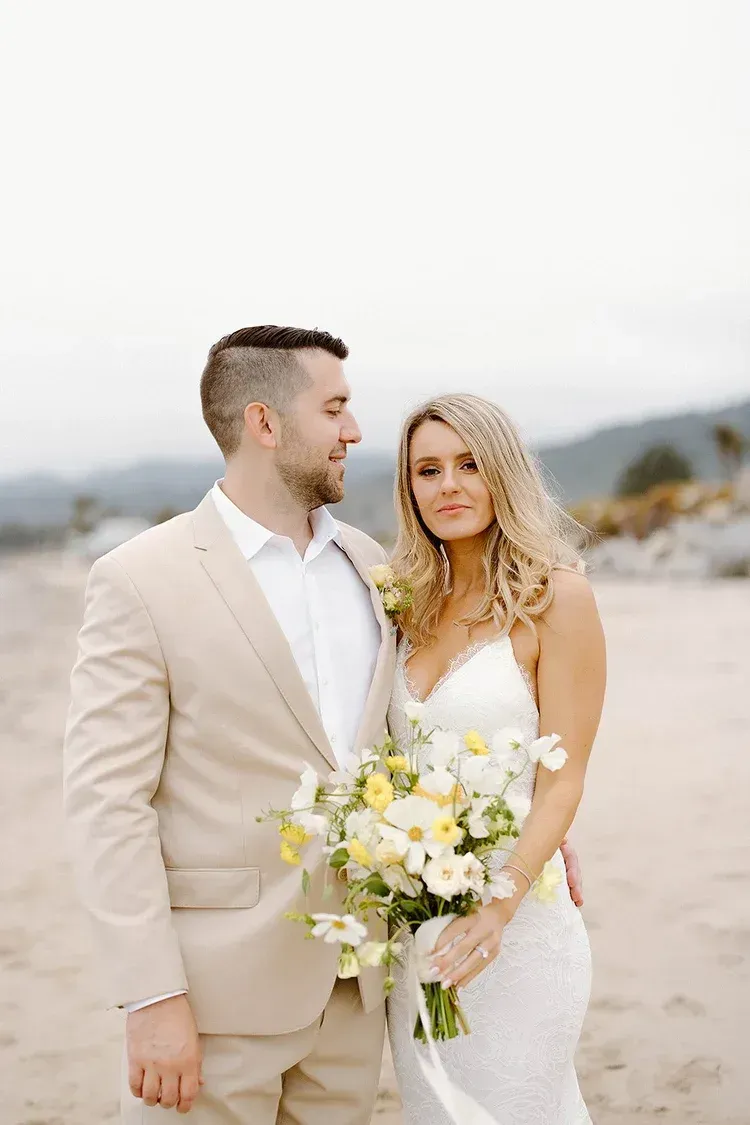 Couple in wedding attire standing on a beach, holding a bouquet, with soft dunes in the background