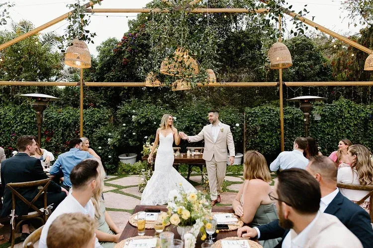Wedding couple holding hands at an outdoor reception under hanging lights, guests seated around long tables