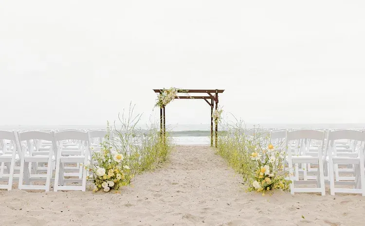 Outdoor wedding aisle with white chairs and floral decorations leading to a wooden arch by the sea